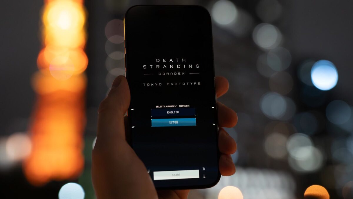 A hand holds a phone showing the Death Stranding Odradek: Tokyo Prototype start screen in front of Tokyo Tower at night